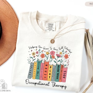 May include: Ivory-colored t-shirt with the text "Helping You Grow To Your Full Potential" and "Occupational Therapy." The design features colorful flowers and books labeled with occupational therapy concepts. A gold necklace is draped on the shirt.