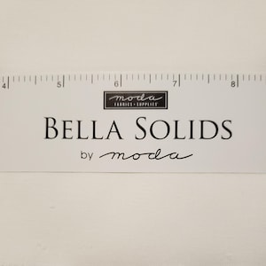 May include: White fabric swatch with a ruler and the text "Bella Solids by moda Fabrics - Supplies" printed on it.
