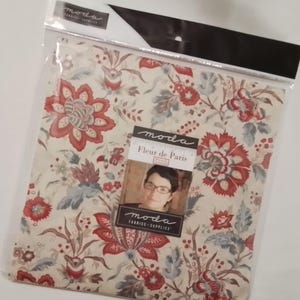 May include: A package of Moda Fabrics fabric featuring a floral pattern in red, cream, and blue. The fabric is in a clear plastic bag with a black and white header. A tag with the text "Fleur de Paris" and a photo is attached.