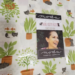 May include: White fabric with a repeating pattern of potted plants, gray cats, and fish bowls with orange fish. A tag reads "HERE KITTY KITTY" and "moda FABRICS + SUPPLIES". The fabric features green, brown, and gray colors.
