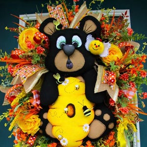 May include: A whimsical door wreath featuring a black bear holding a yellow beehive. The wreath is adorned with orange, yellow, and green floral accents, ribbons, and a smiling bee. The text "Front Door Wreaths" is visible at the bottom.