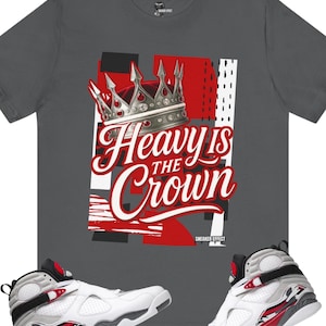 Puede incluir: Camiseta gris oscuro con un gráfico de corona plateada y la frase "Heavy is the Crown" en blanco y rojo. Zapatillas blancas, negras y rojas en la parte inferior de la imagen.
