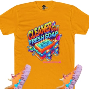 May include: Mustard yellow t-shirt with a graphic that reads "CLEANER THAN FRESH SOAP" and a bar of soap design. The shirt is paired with orange and purple sneakers. The sneakers have a unique, textured sole.
