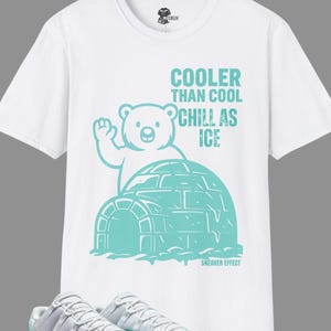 Puede incluir: Camiseta blanca con un gráfico turquesa de un oso saludando desde un iglú, con el texto "Cooler Than Cool Chill As Ice". Debajo, un par de zapatillas blancas y turquesas.