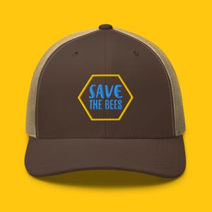 Peut inclure: Un chapeau de camionneur brun et beige avec un hexagone jaune et le texte "SAVE THE BEES" en lettres bleues.