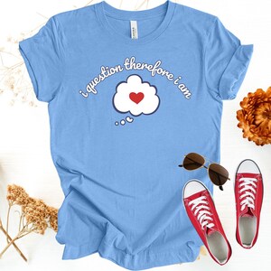 Könnte beinhalten: Hellblaues T-Shirt mit dem Schriftzug "i question therefore i am" und einer Sprechblase mit rotem Herz. Das Bild zeigt auch rote Turnschuhe, eine Sonnenbrille und Trockenblumen.