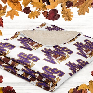 Puede incluir: Manta de forro polar blanca y morada con el texto "Vikes Football" y un gráfico de fútbol americano. La manta tiene un interior suave y afelpado y está doblada. Hojas de otoño rodean la manta.