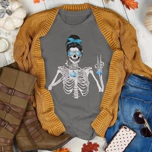 Könnte beinhalten: Ein graues Raglan-Ärmel-T-Shirt mit einem Skelett-Design, das eine Sonnenbrille, ein blaues Stirnband und eine Kaffeetasse mit einem Kürbis trägt. Das Shirt wird mit einer senfgelben Strickjacke, blauen Jeans und braunen Stiefeln kombiniert. Eine Handyhülle und Sonnenbrille sind ebenfalls zu sehen.