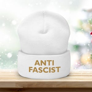 May include: A white beanie hat with the words "ANTI FASCIST" in gold. The hat is made of a soft material and is displayed on a wooden surface. A red star ornament and a Christmas tree are in the background.