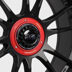 May include: Close-up of a black forged wheel with a red central lock cap. The cap has the OZ Racing logo and the text "LOSEN/REMOVE CENTRALLOCK ANZIEHEN/TIGHTEN FORGED" around the edge. The wheel also has the text "MADE IN ITALY" on the side.