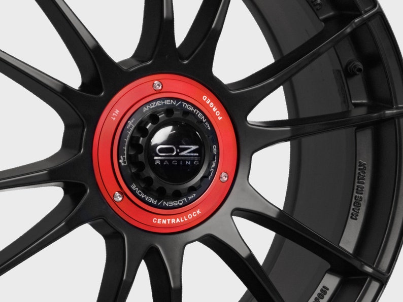 May include: Close-up of a black forged wheel with a red central lock cap. The cap has the OZ Racing logo and the text "LOSEN/REMOVE CENTRALLOCK ANZIEHEN/TIGHTEN FORGED" around the edge. The wheel also has the text "MADE IN ITALY" on the side.