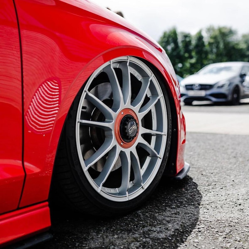 May include: Close-up of a red car with a silver and black wheel. The wheel has a multi-spoke design and a red center cap with the letters "OZ" on it.