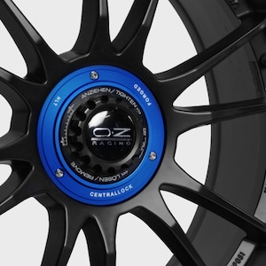 May include: Close-up of a black forged wheel with a blue central lock cap. The cap has the OZ Racing logo and the text "ANZIEHEN/TIGHTEN" and "LOSEN/REMOVE". The wheel also has the text "MADE IN ITALY" on the rim.