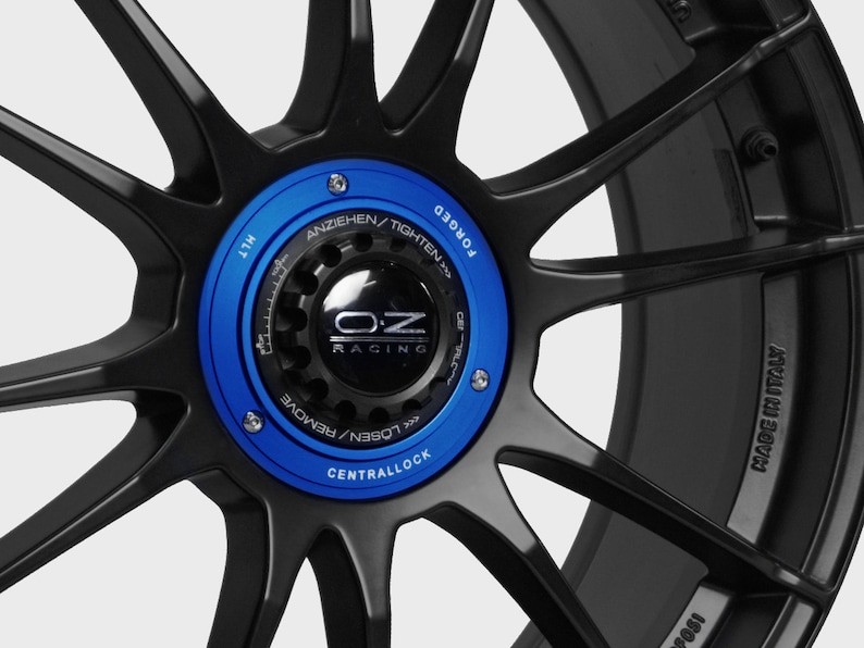 May include: Close-up of a black forged wheel with a blue central lock cap. The cap has the OZ Racing logo and the text "ANZIEHEN/TIGHTEN" and "LOSEN/REMOVE". The wheel also has the text "MADE IN ITALY" on the rim.