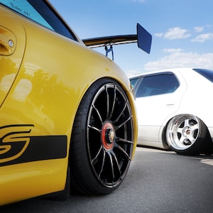 May include: A yellow Porsche 911 with a black rear wing and black wheels. The car has a black and white stripe on the side with the letter "S" in white. The car is parked next to a white sedan.