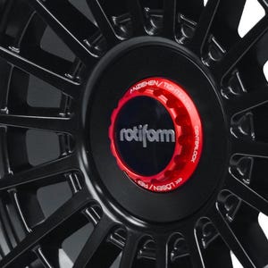 May include: Close-up of a black car wheel with a multi-spoke design. The center cap is black with a red ring and the word "rotiform" in white. The red ring has the text "ANZIEHEN/TIGHTEN" and "CENTERLOCK".