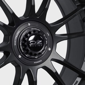 May include: Black forged aluminum car wheel with a silver OZ Racing logo and the text "CENTRALLOCK ANZIEHEN/TIGHTEN >>>>>>> CENTERLOCK" and "MADE IN ITALY".
