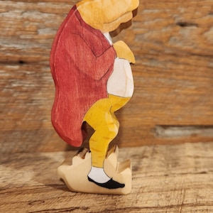 May include: A wooden figurine of a frog wearing a red jacket, yellow pants, and black shoes. The frog is standing on a wooden base.