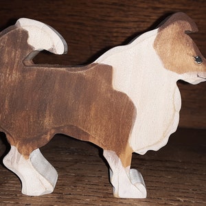 May include: Wooden dog figurine, featuring a brown and white color scheme. The dog is depicted in a standing pose, with its head turned to the side. The figurine is made of wood and has a smooth finish, showcasing the natural grain.