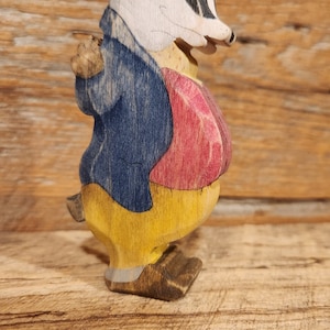 May include: A wooden figurine of a badger wearing a blue jacket, yellow pants, and a red vest. The badger has a white face with black markings.