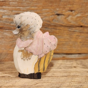May include: A hand-carved wooden figurine of a character with a light brown face, wearing a white bonnet and a pink shawl. The figure has a cream-colored body with yellow and brown striped accents. The figurine is set against a wooden background.