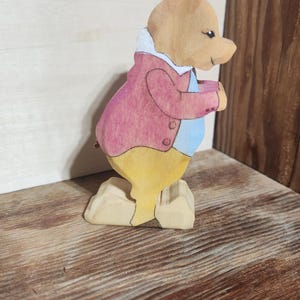 May include: A hand-carved wooden figurine of a rabbit standing on a base. The rabbit is painted with a light brown head, a pink jacket with a white collar, a light blue shirt, and yellow trousers. The figure is shown in profile facing to the right.