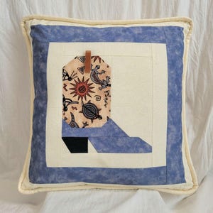 May include: Square decorative pillow with a Southwestern design. The pillow features a blue border, a cream-colored center, and a boot-shaped applique with Native American-inspired patterns. A leather tag is attached to the boot.