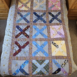May include: A rectangular quilt featuring a grid of square blocks, each containing a central X-shaped fabric applique. The quilt is constructed from various patterned fabrics in blue, yellow, pink, and plaid, joined by brown floral sashing with yellow yarn ties.