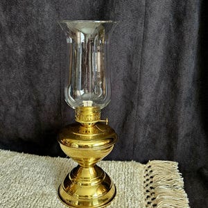 May include: A vintage-style oil lamp with a clear glass chimney and a polished brass base. The lamp features a round reservoir and a decorative, ornate base. The glass chimney is flared at the top. The lamp is sitting on a beige rug.