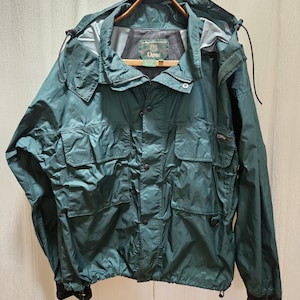 May include: A dark green hooded jacket featuring a front zipper closure with a snap-button placket, multiple utility pockets, and adjustable cuffs. The interior label displays the Orvis brand name. The jacket includes drawstrings at the hood and hem.