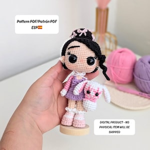 Könnte beinhalten: Handgemachte Häkelpuppe mit schwarzen Haaren, lila Kleid und rosa Schuhen, die ein weißes und rosa Häschen hält. Die Puppe steht auf einem Holzsockel. Der Text auf dem Bild lautet "Pattern PDF/Patrón PDF ESP" und "DIGITAL PRODUCT - NO PHYSICAL ITEM WILL BE SHIPPED."