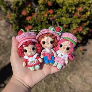 May include: Three handmade crochet dolls, each with a strawberry theme. The dolls have pink hats with strawberry accents, and wear dresses or outfits in red, pink, and white. The dolls are held in a hand, with a blurred green and red background.