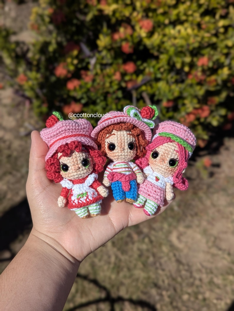 May include: Three handmade crochet dolls, each with a strawberry theme. The dolls have pink hats with strawberry accents, and wear dresses or outfits in red, pink, and white. The dolls are held in a hand, with a blurred green and red background.