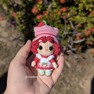 May include: A handmade crochet doll of a character with red hair, a pink hat with a green strawberry, and a red dress with a white apron. The doll is held in a hand. The doll's eyes are black. The doll is approximately 4 inches tall.