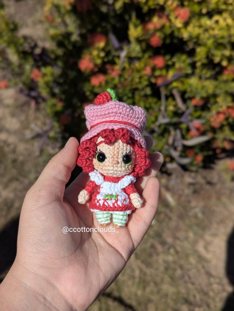 May include: A handmade crochet doll of a character with red hair, a pink hat with a green strawberry, and a red dress with a white apron. The doll is held in a hand. The doll's eyes are black. The doll is approximately 4 inches tall.