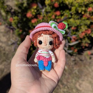 May include: A handmade crochet doll with a pink hat adorned with a red flower and green leaves. The doll has brown hair, a striped shirt, blue pants, and a pink scarf. The doll is held in a hand.