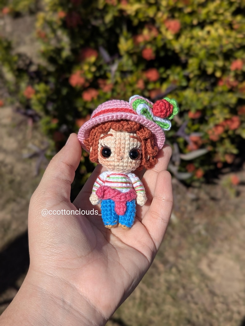 May include: A handmade crochet doll with a pink hat adorned with a red flower and green leaves. The doll has brown hair, a striped shirt, blue pants, and a pink scarf. The doll is held in a hand.