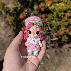 May include: A handmade crochet doll resembling a strawberry. The doll has pink hair, a pink dress, and a green and white striped hat and socks. The doll is held in a hand against a blurred background of green foliage. The doll has large black eyes.