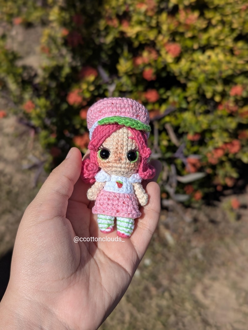 May include: A handmade crochet doll resembling a strawberry. The doll has pink hair, a pink dress, and a green and white striped hat and socks. The doll is held in a hand against a blurred background of green foliage. The doll has large black eyes.