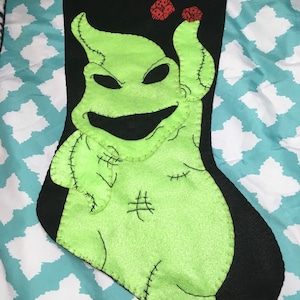 May include: A green and black Christmas stocking with a stitched-on image of the character Oogie Boogie from the movie The Nightmare Before Christmas. The stocking has a black faux fur cuff and two red ornaments.