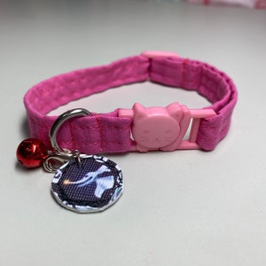 May include: Pink cat collar with a cat-shaped buckle and a silver charm with a red bell. The charm has a black and white image of a cat.