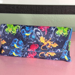 Peut inclure: Une pochette en tissu noir avec un imprimé de dragons colorés. La pochette a une fermeture à bouton rouge et une doublure bleu foncé.