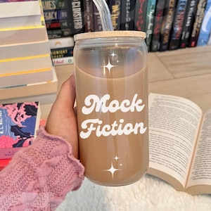 May include: A glass tumbler with a brown beverage and a bamboo lid. The tumbler has a white design that says "Mock Fiction" with stars on either side of the text. A clear glass straw is in the tumbler.