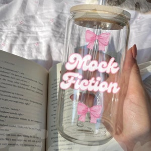 May include: A clear glass tumbler with a wooden lid and a glass straw. The tumbler has two pink bows on it and the text "Mock Fiction" in white letters with a retro font. The tumbler is being held by a hand.