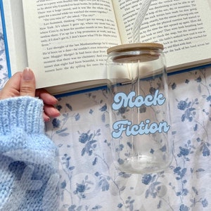May include: A clear glass jar with a wooden lid and a blue and white sticker that says "Mock Fiction". The jar is sitting on a white surface with a blue floral pattern.