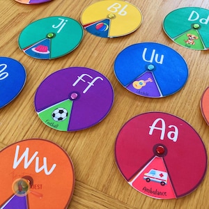 May include: A set of colorful, circular learning aids for children. Each circle features a letter of the alphabet, a picture, and a word. The letters are Aa, Bb, Dd, Ff, Ji, Uu, and Ww. The pictures include a ball, a dog, a football, a jelly, a ukulele, and an ambulance.