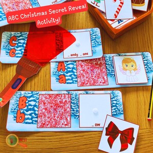 Puede incluir: Un juego educativo de Navidad para niños. El juego incluye una lupa roja, una tarjeta azul y blanca con la letra "B" y la palabra "_OW", y una tarjeta separada con un lazo rojo.