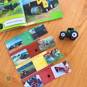 May include: A set of colorful cards with pictures of farm equipment and the words "Trailer", "Tedder", "Plough/Plow", and "Snowblower" printed on them. The cards are laid out on a wooden surface with a toy tractor in the upper right corner.