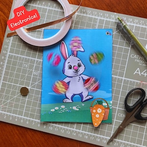 May include: A craft project setup with a cartoon bunny juggling Easter eggs on a blue background. The image includes copper tape, scissors, a pen, and the text "DIY Electronics!"