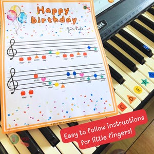 May include: A colorful music sheet with the words "Happy Birthday for Kids" and musical notes. The sheet has colorful shapes and confetti, with the text "Easy to follow instructions for little fingers!" on a red banner. The sheet is placed on a piano keyboard.